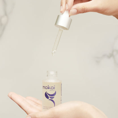 A close-up of a skincare dropper bottle with a clear glass dropper being held above an open hand, with a drop of clear serum about to be transferred. The bottle has a beige label with a purple logo and the brand name 'makai' visible.
